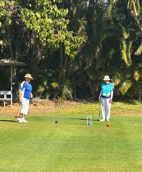 Two women in blue shirts and hats play croquet on a sunny lawn, with one preparing to strike a ball near a wicket