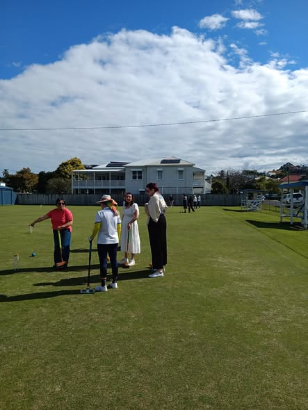 Four people on a croquet lawn, one preparing to hit a ball with a mallet, with croquet wickets and a building in the background