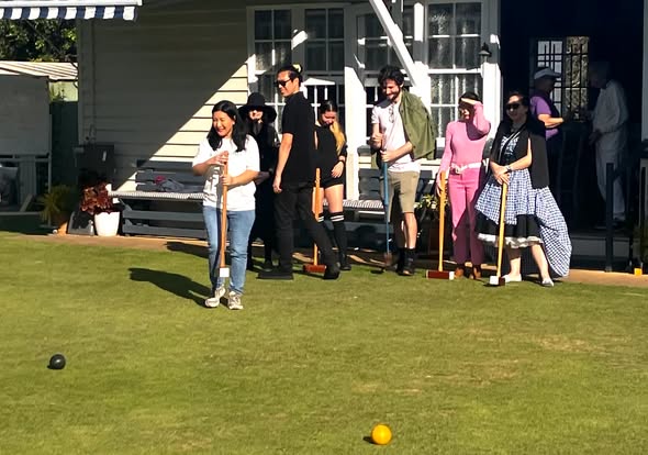 A group plays croquet on a sunny lawn, with players holding mallets and aiming at balls near a house