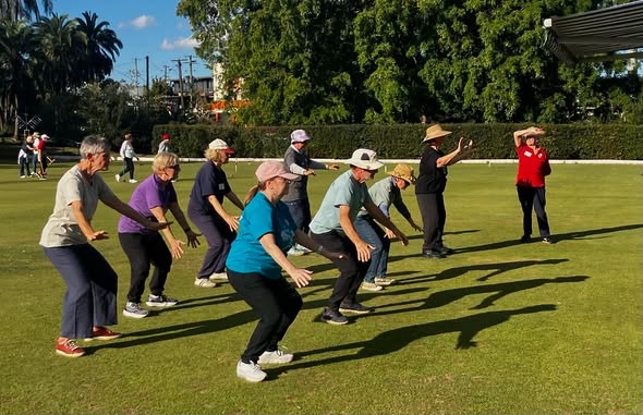A group of people in croquet attire practice a synchronized warm-up on a sunny lawn, with one person in red demonstrating the movement
