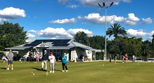 Players enjoy a sunny day of croquet on a well-maintained lawn with a solar-powered clubhouse in the background