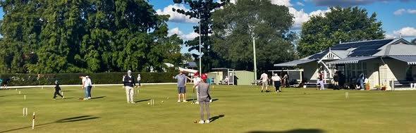Players compete in a croquet match on a sunny day at a club, with mallets, wickets, and a clubhouse visible