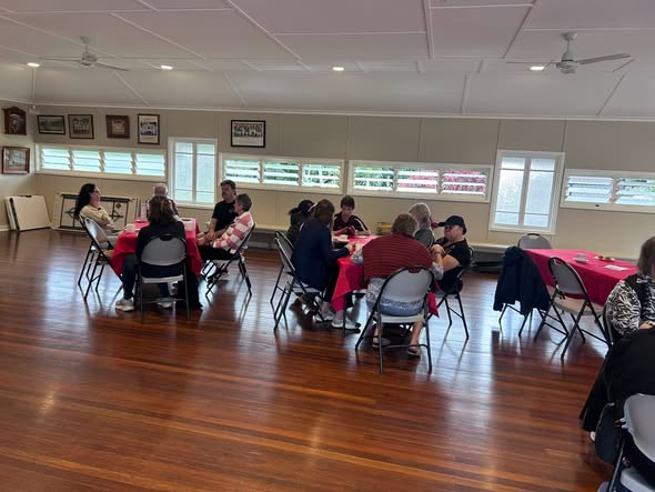People seated at tables with red tablecloths inside a croquet club hall, likely during a meeting or social event