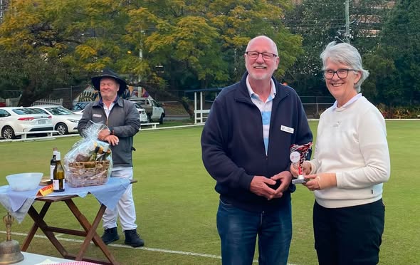 Two people pose with a trophy on a croquet lawn, with another person and a prize table in the background