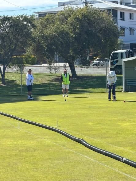 Three people play croquet on a sunny lawn, with one player preparing to strike a ball near a wicket, while others observe