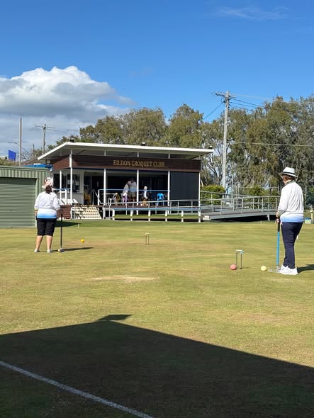 Two players at the Eildon Croquet Club play on a sunny day, with one preparing to hit a ball with a mallet near a wicket