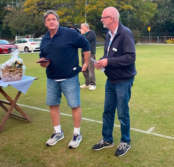Two men stand on a croquet lawn, one holding a clipboard, near a prize basket, during a croquet event
