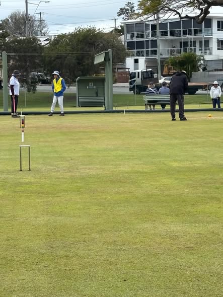 Players compete in a croquet match on a grass court, with one person in a yellow vest preparing to hit a ball near a wicket