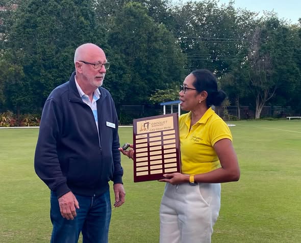 A woman in a yellow polo holds a croquet trophy plaque while speaking with a man on a croquet lawn