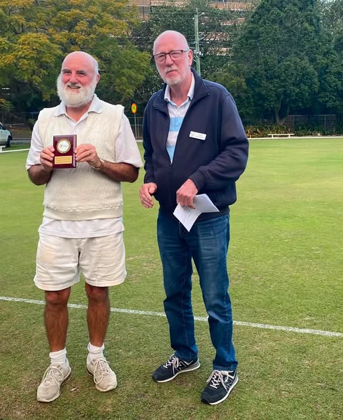 Two men stand on a croquet lawn; one holds a trophy, the other a paper, suggesting a competition award ceremony