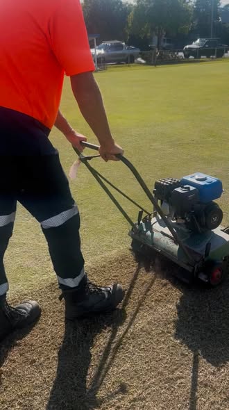 A groundskeeper in an orange shirt operates a motorized lawn roller on a croquet lawn, compacting the surface for play