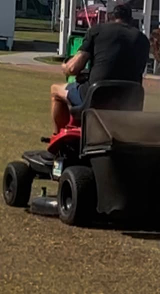 A man operates a red riding lawnmower on a grassy croquet field, with a green croquet ball visible in the background