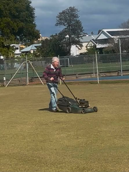 An older man mows a grassy croquet lawn with a push mower, with croquet wickets and a fence visible in the background