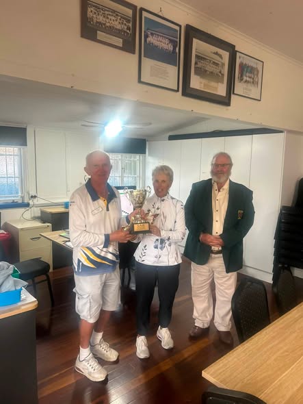 Three people in croquet attire stand indoors, holding a trophy, with framed photos on the wall behind them