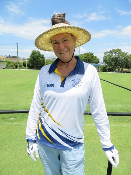 A woman in a croquet uniform and straw hat smiles on a sunny lawn, holding a croquet mallet