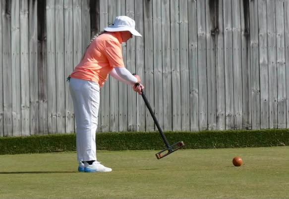 A player in a pink shirt and white hat prepares to strike a red croquet ball with a mallet on a grass court