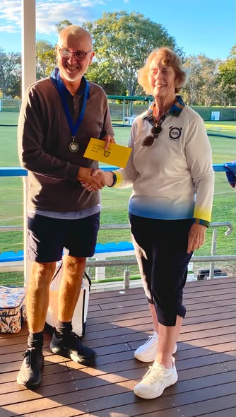A man and woman shake hands on a deck, with the man holding a yellow certificate and a medal, on a croquet lawn under sunny skies