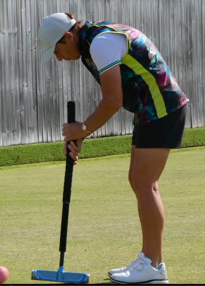 A woman in a colorful shirt and white cap prepares to strike a croquet ball with a mallet on a grassy court