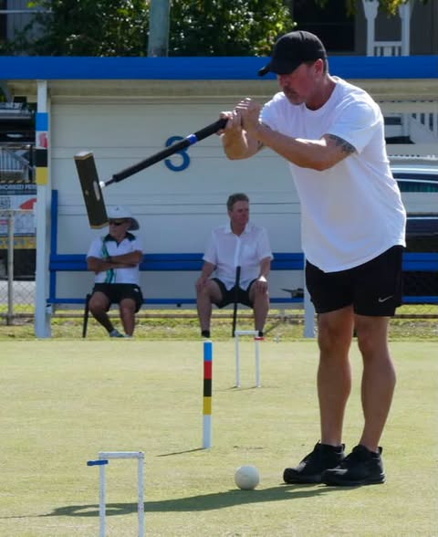 A man in a white shirt and black cap swings a croquet mallet at a ball on a grass court, with two spectators watching from a bench