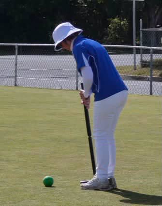 A person in a blue shirt and white hat prepares to hit a green croquet ball with a mallet on a grass court