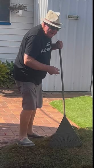 A man in a hat and sunglasses uses a rake on a brick patio near a croquet lawn